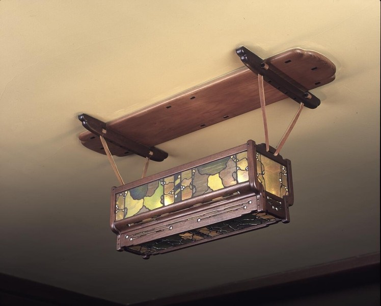 Elongated rectangular chandelier with substantial wooden crossbeam support and decorative end brackets. The suspended fixture features amber stained glass panels with organic vine patterns in dark metal frame, exemplifying Greene and Greene's integration of Japanese-influenced design with Arts and Crafts principles.