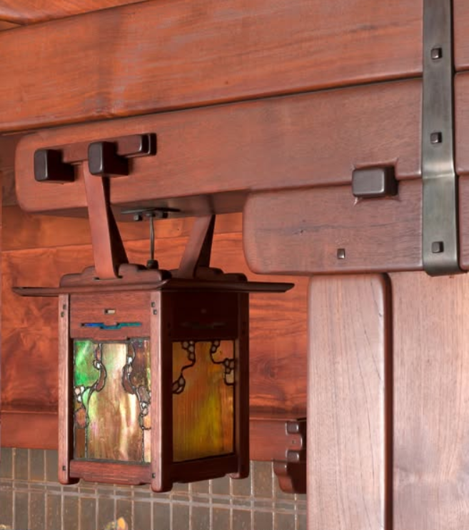 Inglenook lantern suspended by leather straps from wooden brackets with decorative metal hardware. Features art glass panels in amber, green, and iridescent tones within dark wood frame. Mounted against Honduras mahogany paneling with visible pegs and metal strap details, exemplifying Greene and Greene's innovative use of materials.