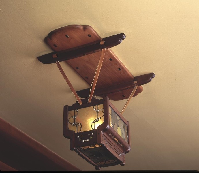 Compact chandelier with wooden crossbeam canopy and suspended rectangular lantern body. Features metal frame with decorative cutouts and amber glass panels, showing Arts and Crafts joinery details including visible pegs and hardware, suspended by copper-toned rods against cream-colored ceiling.