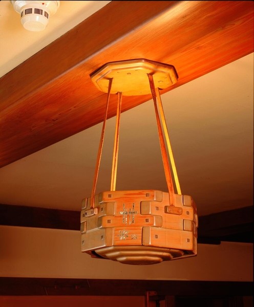 Cylindrical wooden ceiling pendant with geometric Arts and Crafts design, suspended by four rods from an octagonal canopy. The fixture features decorative cross motifs and square peg details in natural wood finish, illuminated warmly and positioned beneath diagonal exposed ceiling beams.