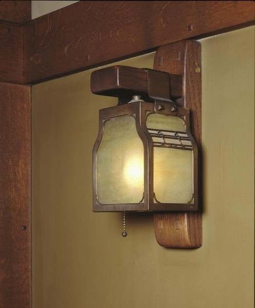 Arts and Crafts wall sconce with carved wooden backplate and bracket supporting a square lantern with amber glass panels. Features metal frame with decorative cutouts, pull-chain switch, and exposed joinery details mounted against pale yellow wall beside dark wood beam.
