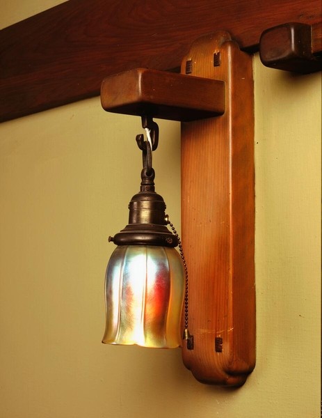 Wall-mounted sconce with wooden backplate and bracket supporting an iridescent art glass shade in bell form. The fixture displays rainbow-like color effects in amber and green tones, with metal hardware and pull-chain switch, mounted against pale yellow wall beside dark wood trim.