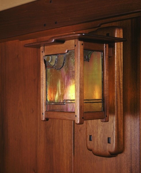 Dining room wall sconce with substantial wooden backplate featuring Arts and Crafts joinery. Rectangular lantern with iridescent art glass panels in amber and pink tones, set within dark metal frame with decorative hardware, mounted on rich wood-paneled wall.