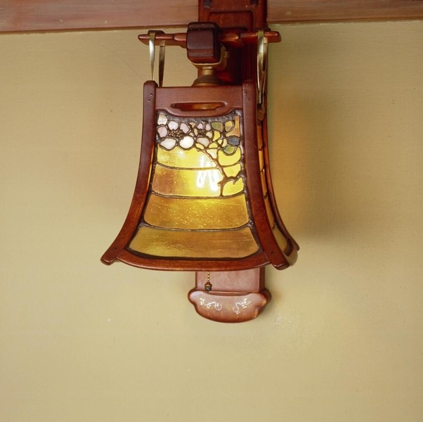 Exceptional wall sconce crafted from Honduras mahogany with silver inlay details, featuring curved organic backplate. Art glass shade displays wisteria motif in purple, yellow, and green leaded glass panels, exemplifying Greene and Greene's masterful integration of decorative arts.