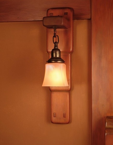 Master bedroom wall sconce with elongated wooden backplate and chain-suspended bell-shaped shade in frosted amber glass. Features brass or bronze hardware with decorative details, mounted on pale yellow wall beside natural wood trim in warm lighting.