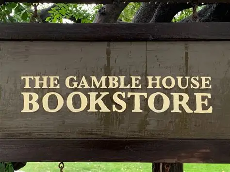 Wooden sign with gold serif lettering reading "THE GAMBLE HOUSE BOOKSTORE" mounted on dark stained timber beam. Simple, elegant design consistent with Arts and Crafts aesthetic, photographed outdoors with foliage visible in background.