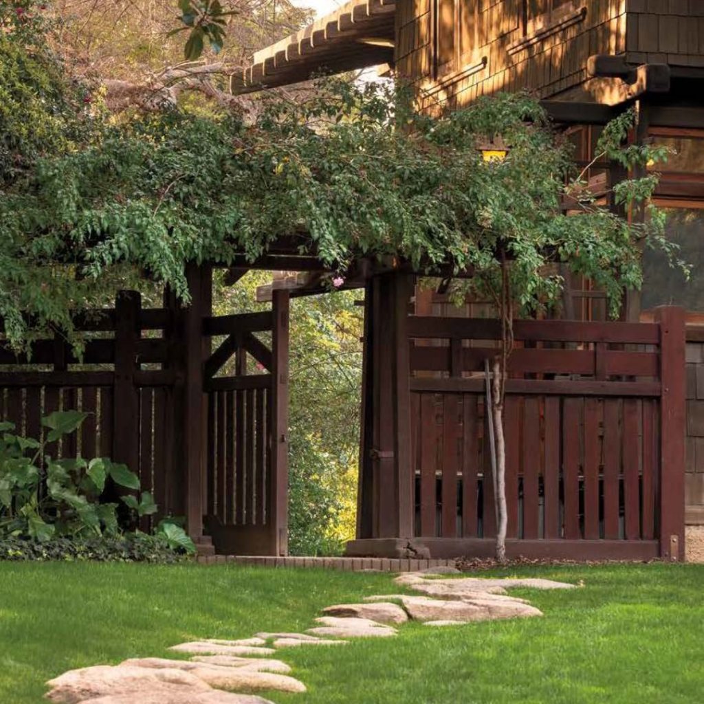 Irregular stepping stones set in manicured lawn leading to dark wood gate structure with vertical slat design. Craftsman-style architecture visible with exposed timber beams, shingled walls, and mature landscaping including ornamental trees creating dappled shade over the garden path.