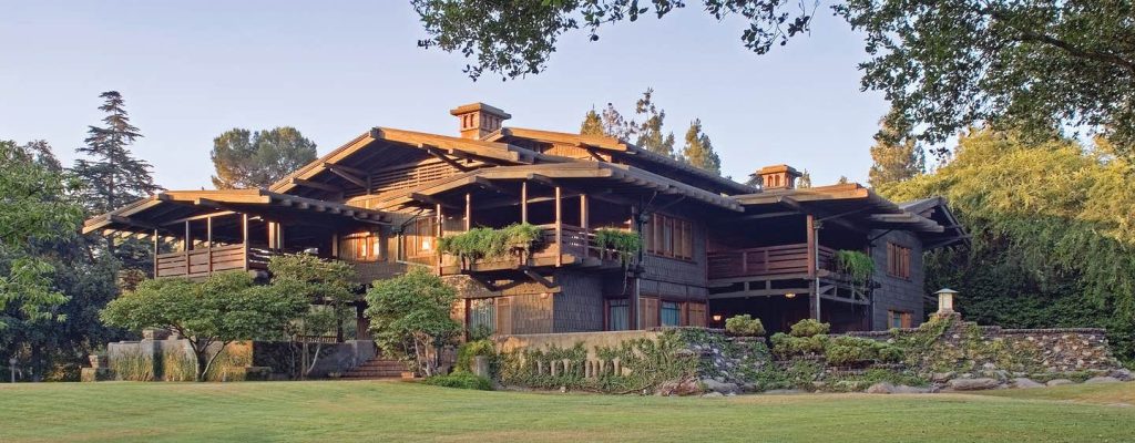 Full northwest facade view of the Gamble House at golden hour showing the complete composition with symmetrical sleeping porches flanking the central entrance, central brick chimney with decorative cupola topped by a flag, multiple low-pitched gabled roofs with exposed rafter tails and decorative brackets, weathered wood shingle siding, terraced clinker brick retaining walls with concrete planters containing spiky yucca plants and ornamental grasses, Arts and Crafts lanterns at the entry, and mature eucalyptus and oak trees framing the property, with warm sunset light illuminating the teak woodwork and creating long shadows across the manicured lawn