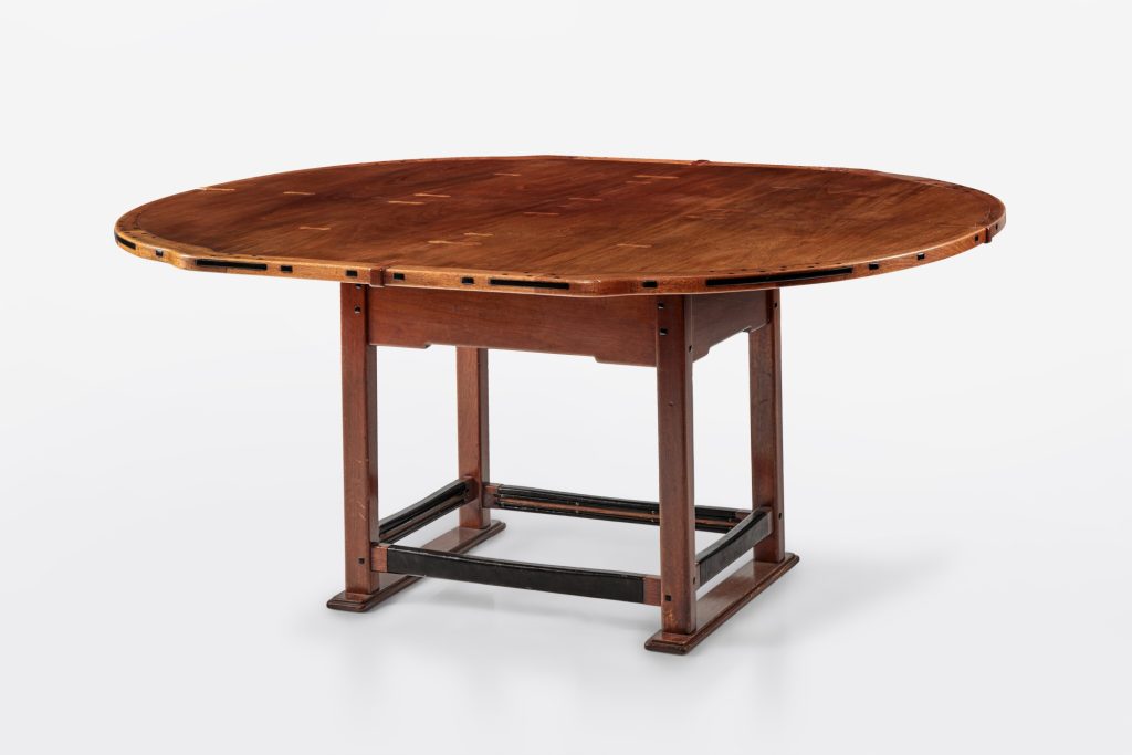 Round dining table with ebony-pegged edge, butterfly inlay on top, and metal stretchers connecting sturdy pedestal base.