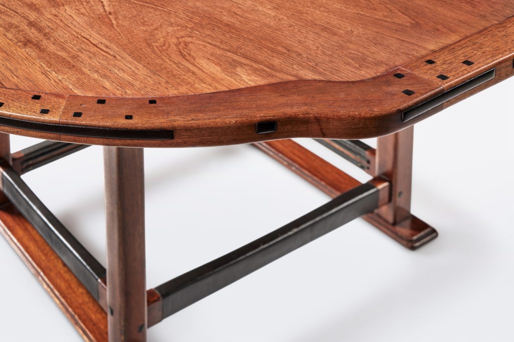 Dining table edge detail with ebony pegs along curved rim, metal stretchers, and exposed joinery characteristic of Greene design.