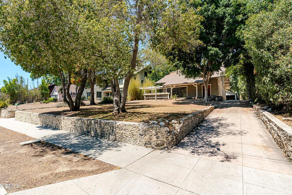 Sidewalk view of Francis F. Rowland house with decorative river rock retaining walls, mature trees, Craftsman bungalow, and white timber pergola