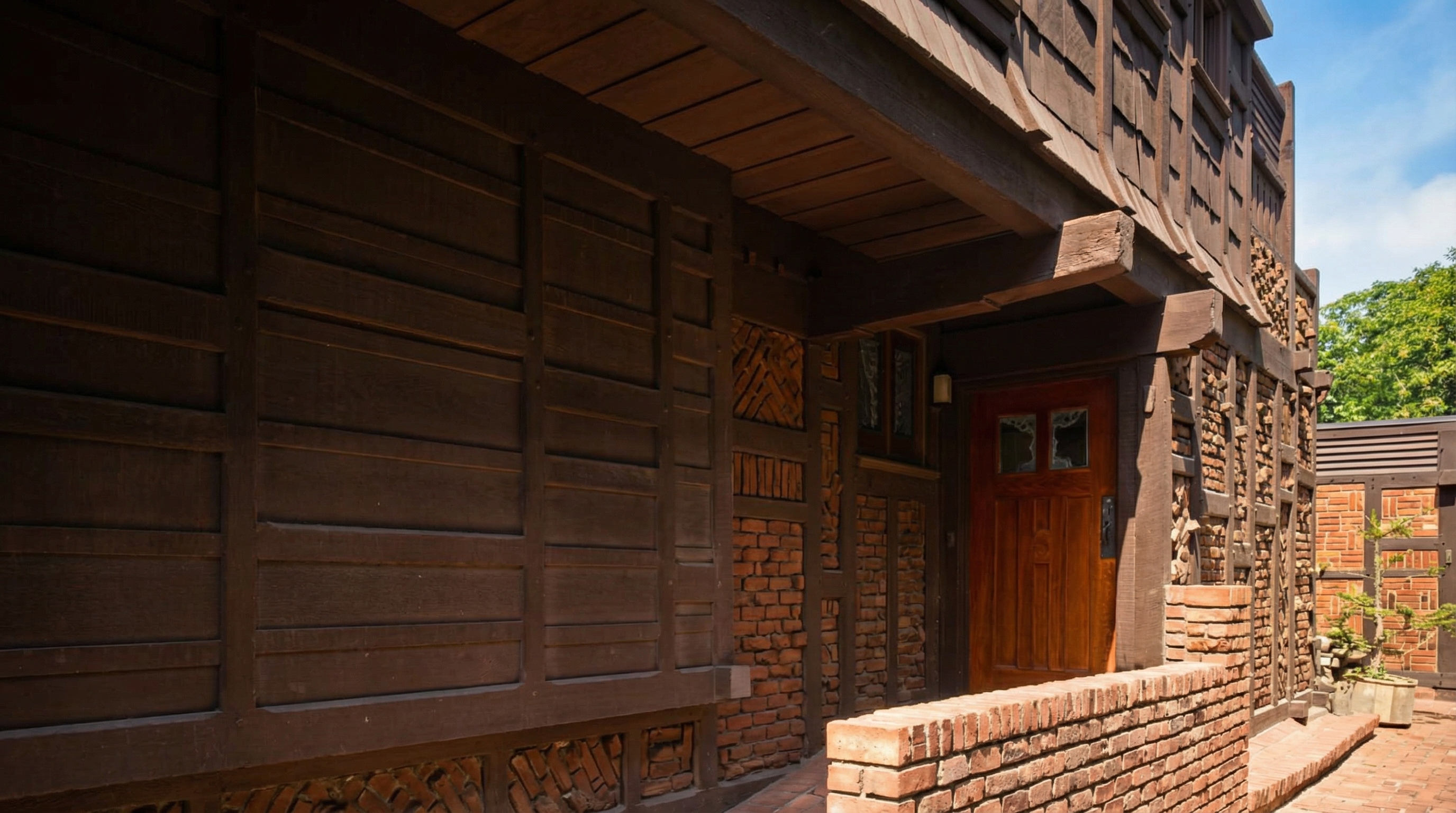 Close view of the entrance walkway at the Adelaide Tichenor House, with redwood board-and-batten walls, clinker-brick detailing, deep timber eaves, and a wood front door with glass lights reached by a brick path