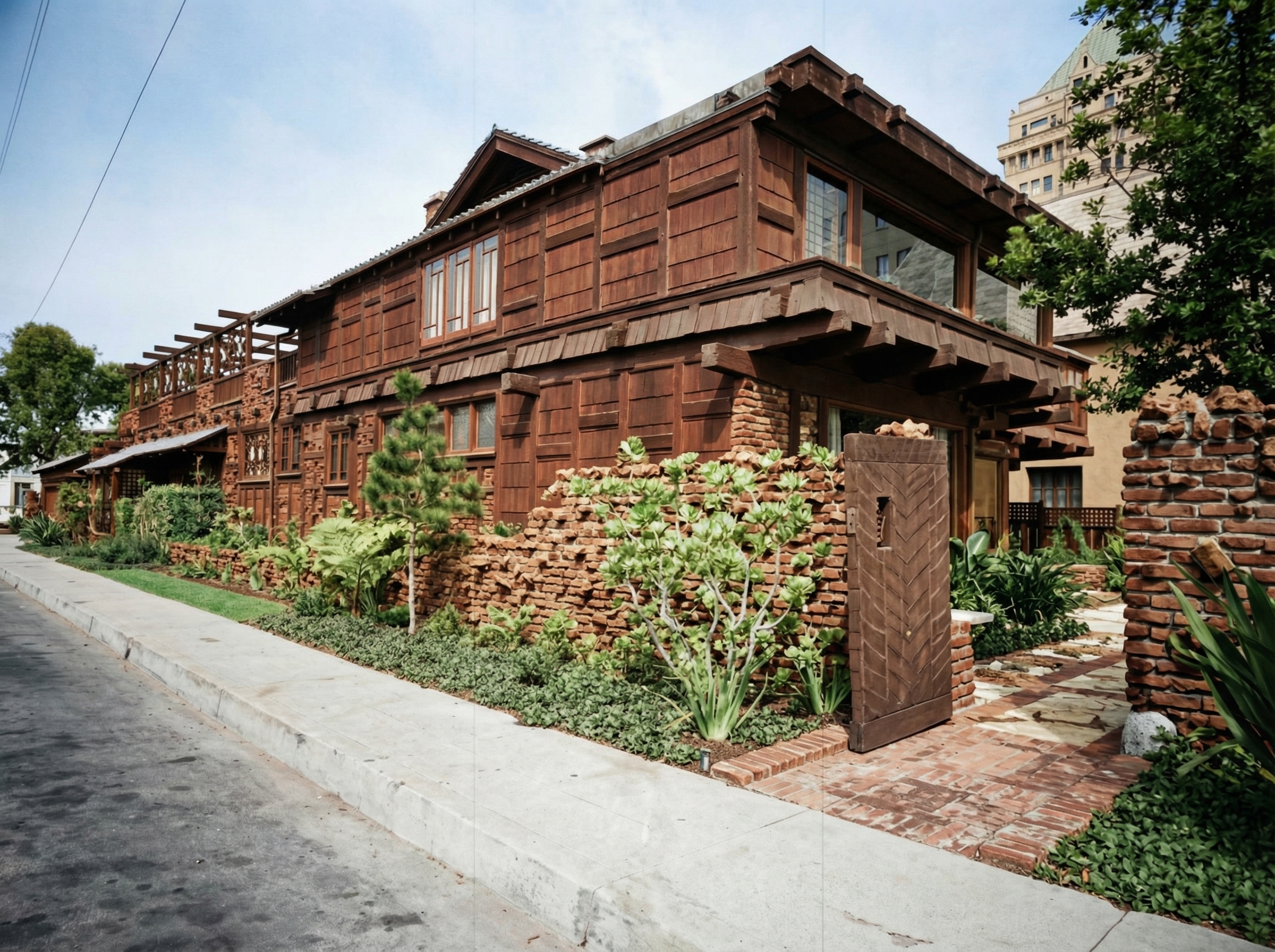 Street view of the Adelaide Tichenor House in Long Beach, a two-story dark-shingled Craftsman house with broad eaves and exposed rafters, a clinker-brick garden wall along the sidewalk, and a wooden herringbone-pattern garden gate set into a brick pier