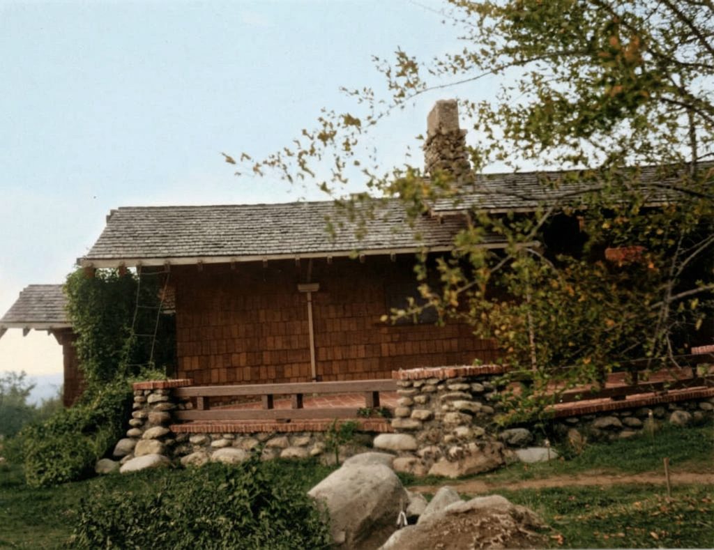 Historic photograph of east terrace with river rock foundation, wood shingle walls, and massive stone chimney amid natural landscaping.