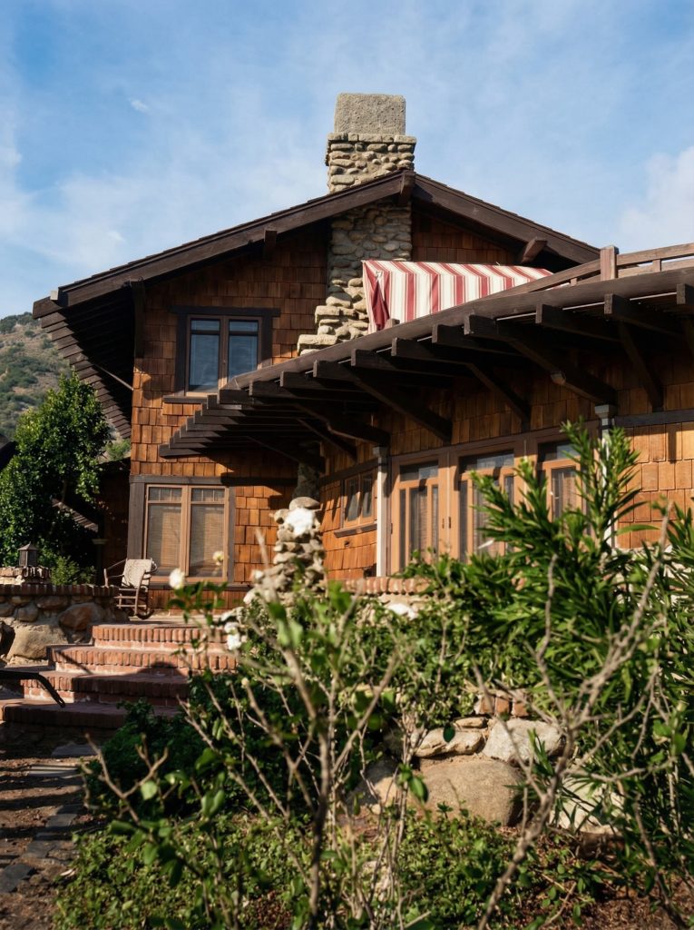 Vintage front view showing west entrance with massive river rock chimney, deep eaves, and naturalistic plantings framing the house.
