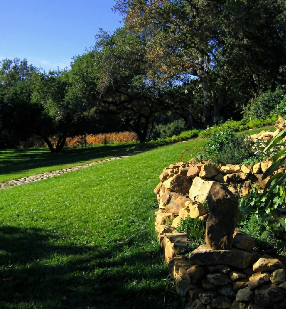 Landscaping view of The Charles M. Pratt House showing garden beds, stone retaining walls, and mature specimen trees