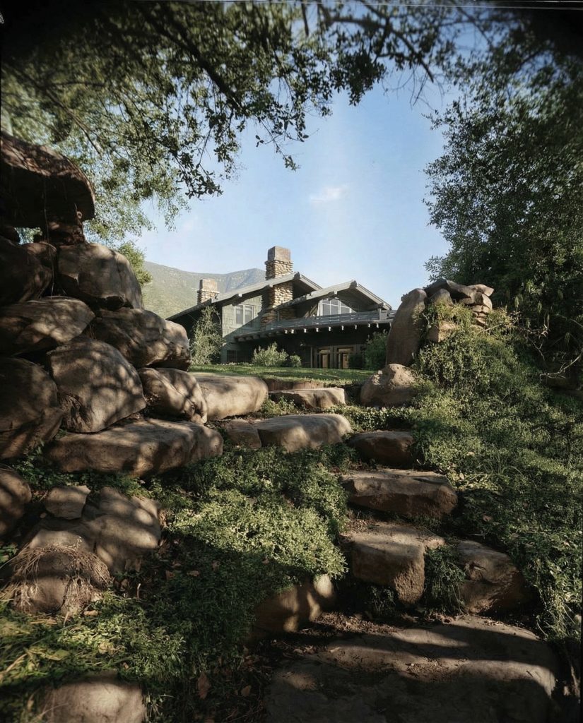 Colorized distant walkway view of The Charles M. Pratt House showing stone paths, gardens, and Craftsman architecture