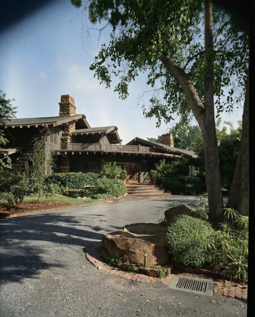 Colorized front entrance driveway of The Charles M. Pratt House showing stone walls, mature trees, and approach path