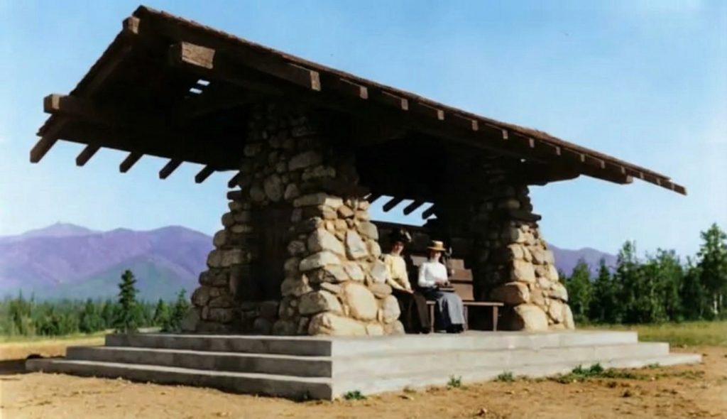 Angled view of the stone and timber pavilion with visitors seated inside, mountains visible in background