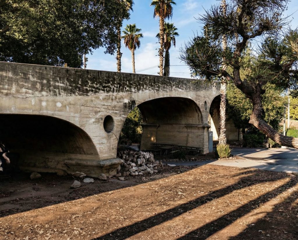 Ground-level view of the Oaklawn Bridge showing its sweeping reinforced concrete arches and weathered patina amid lush vegetation.