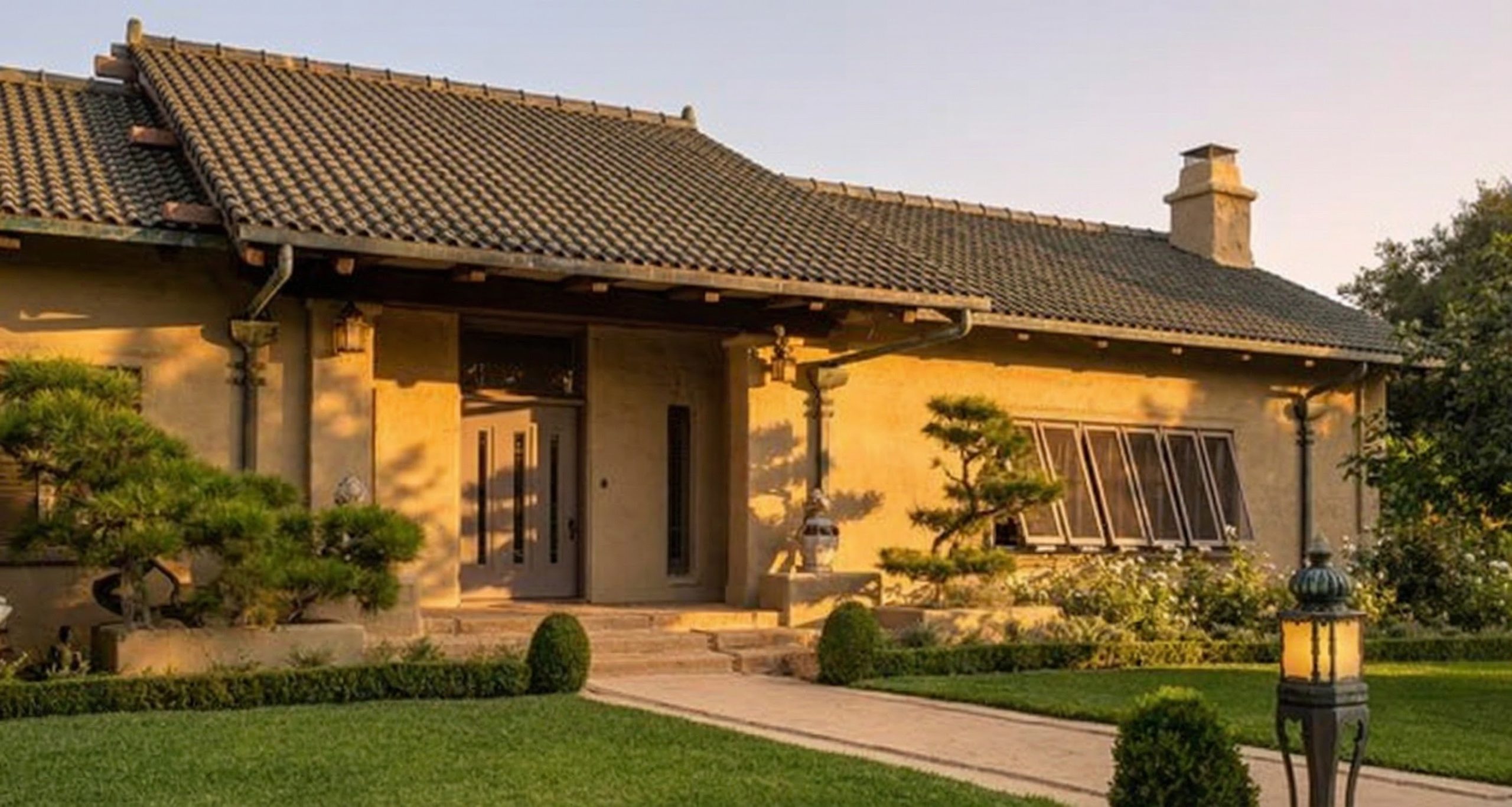 Contemporary street view of the Cordelia A. Culbertson House in Pasadena, a long low building with cream-colored troweled gunite walls and a low red and green tile roof set behind a garden wall, with no exposed timber and no shingled bungalow roof