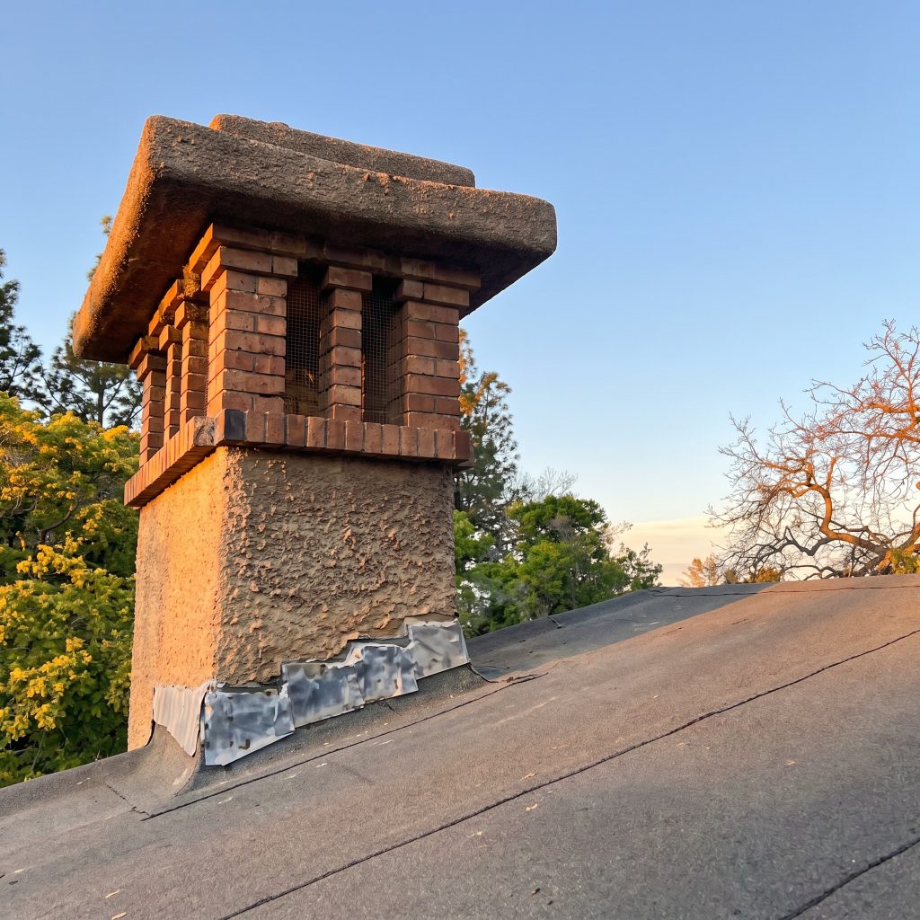 Photograph of the Gamble House chimney cap by Greene and Greene, taken from the roof at sunset, showing a small architectural pavilion at the top of a plain stucco shaft — four corner piers of unclinkered red brick supporting a substantial poured-concrete cap with deeply expressed eaves, the flue openings between the piers fitted with screening, with sky and treetops in the background and the dark roof slope falling away below