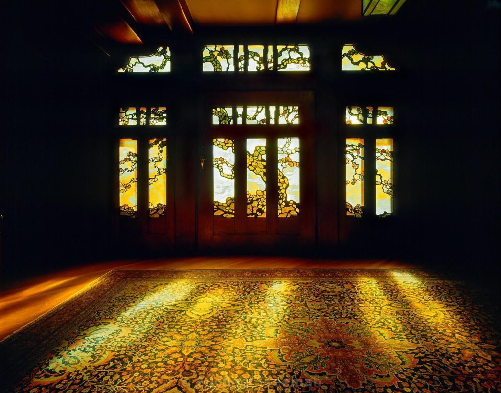 Dramatic interior photograph of the Gamble House front doors seen from the entry hall, showing the leaded art glass by Emil Lange depicting a Japanese black pine silhouetted against backlit amber and white glass, with warm light cast across the original Persian rug and teak floor