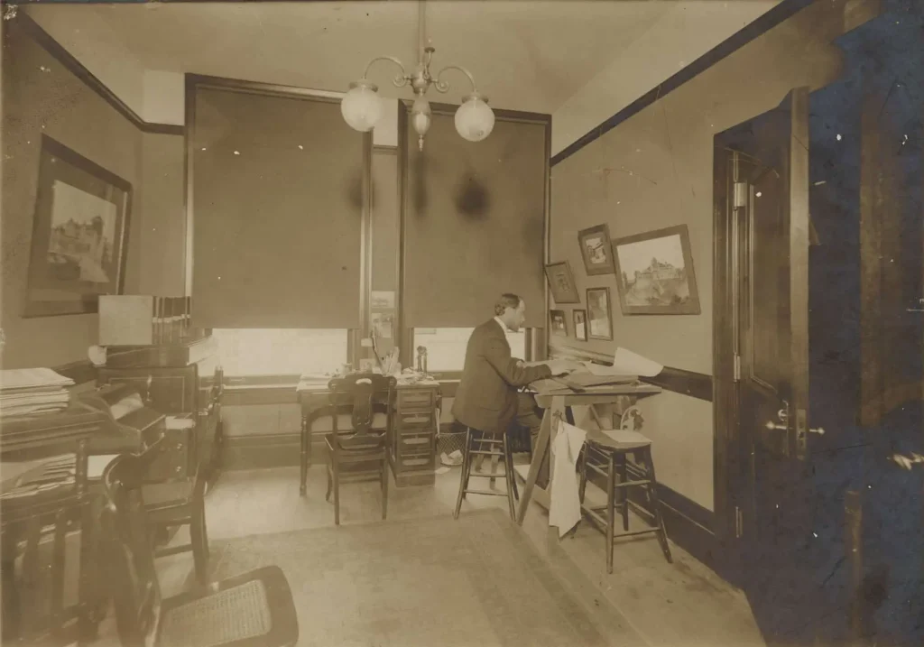 Sepia photograph of architect Henry Greene seated at a drawing board in the Greene and Greene architectural office in Pasadena, California, circa early 1900s, with architectural sketches and paintings visible on the walls