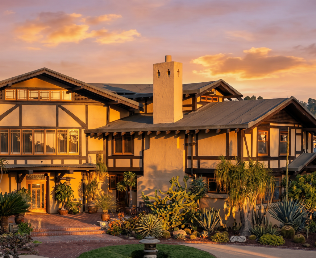 Photograph of the Laurabelle A. Robinson House by Greene and Greene at sunset, showing the half-timbered upper stories and stucco walls with multiple gabled wings centered on a tall stuccoed chimney with iron anchor plates, surrounded by drought-tolerant plantings and a brick courtyard