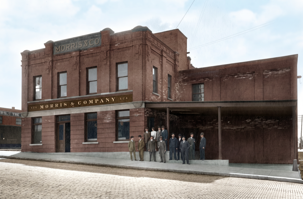 Colorized historic photograph of the Morris and Company building, a two-story brick commercial structure with the firm's name displayed prominently across the façade, with a group of employees standing on the sidewalk in front, photographed in the early twentieth century