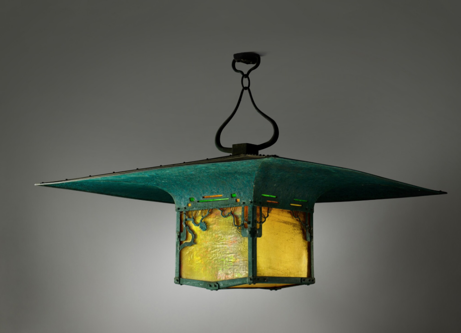 A Greene and Greene hanging lantern from the Blacker House photographed alone against a plain wall, a flaring patinated-copper roof above a box of amber leaded glass.