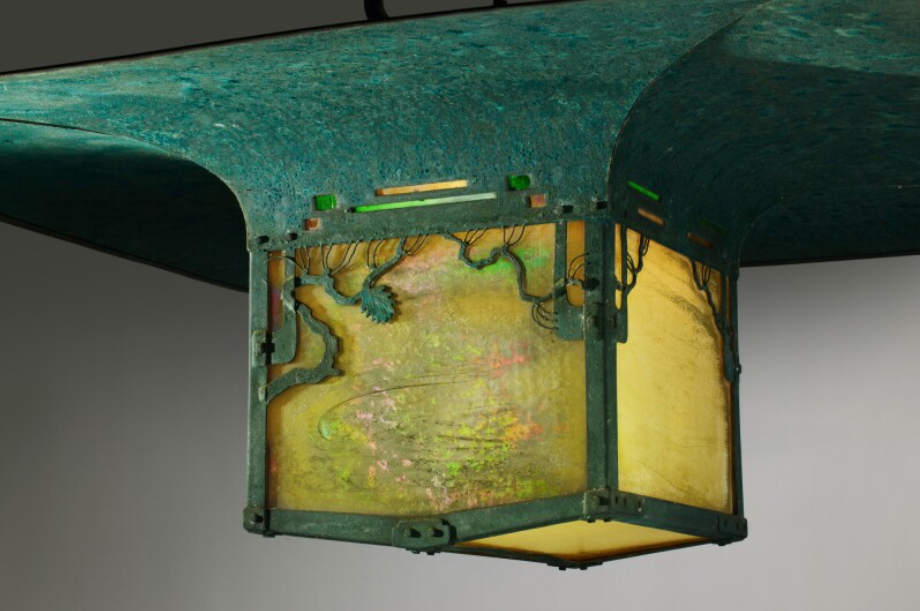 Close-up museum photograph of the Blacker House pagoda lantern showing the verdigris copper roof, the amber leaded art glass panels by Emil Lange filtering warm yellow light, the dark bronze openwork frame, and applied decorative bronze pine branches at the upper border with small inset chips of green and amber glass