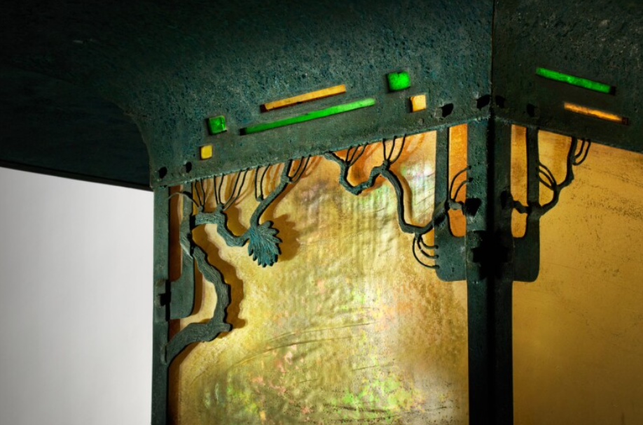 Extreme close-up museum photograph of the bronze openwork pine branch detail at the upper register of the Blacker House porte-cochère lantern, showing the curving bronze branch laid across amber Lange art glass with individual needle clusters articulated as small dimensional sprays, with green and amber glass chips inset above