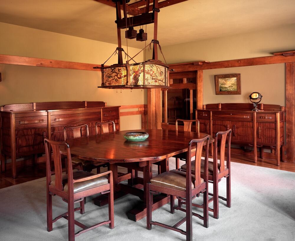 Photograph of the Laurabelle A. Robinson House dining room as installed at the Huntington Library in San Marino, California, showing the original mahogany table and chairs, twin built-in sideboards with copper hardware, the iconic Robinson House art glass chandelier, and the integrated mahogany paneling — preserved as the most important installation of American Arts and Crafts design in any museum