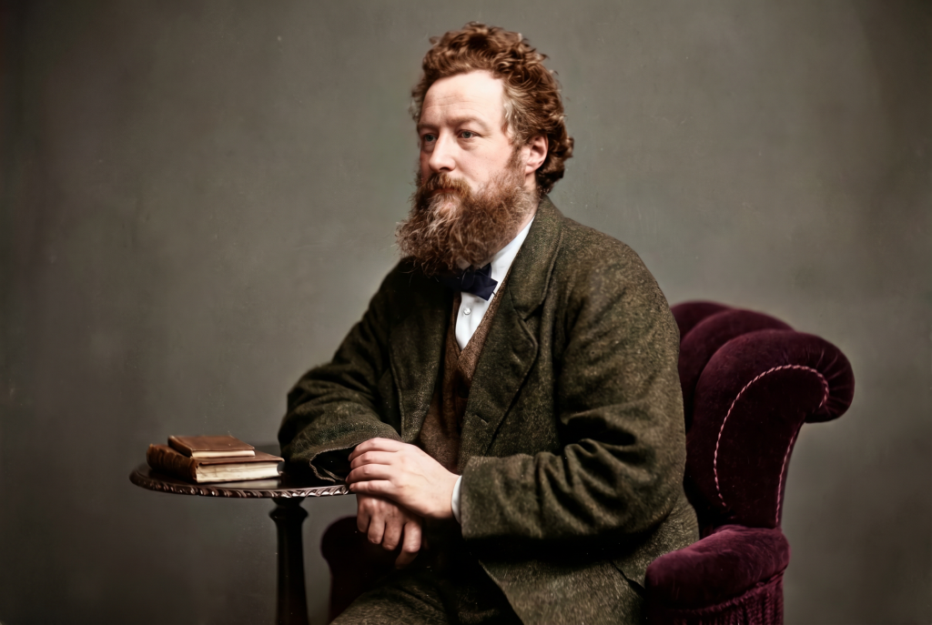 Colorized historic portrait of William Morris seated in a velvet armchair beside a small table holding two books, wearing a tweed jacket and bow tie, his characteristic full beard and intense expression caught in a studio photograph from the 1870s — the founder of the Arts and Crafts movement and author of the contradiction the movement could not resolve