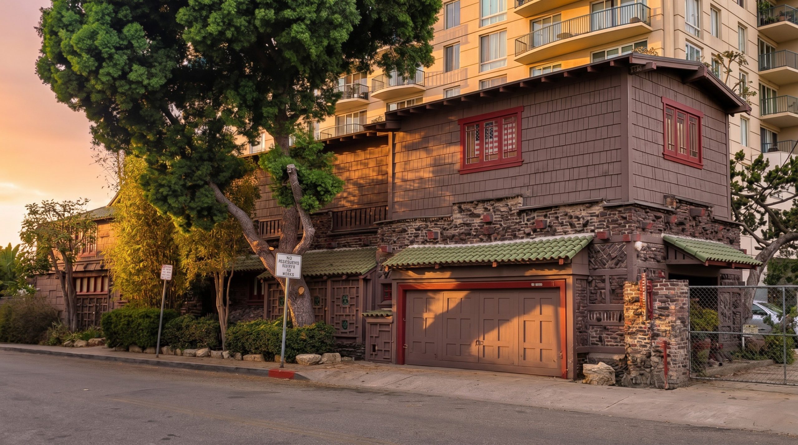 Contemporary photograph of the Adelaide M. Tichenor House as it stands today at 852 East Ocean Boulevard in Long Beach, showing the altered brown-shingle residence with red trim, stone-faced base, attached garage with green-tile roof, mature trees overhead, and a modern high-rise residential tower rising directly behind the property at sunset