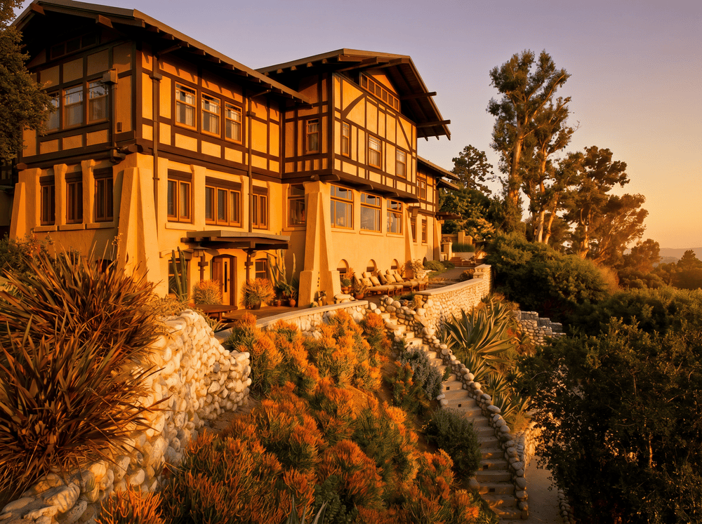 Sunset photograph of the Laurabelle A. Robinson House in Pasadena showing the canyon-facing elevation glowing in golden light, with the stucco walls illuminated, half-timbering reading dark against the sky, river-stone retaining walls stepping down through the arroyo-edge garden, and tapered buttresses anchoring the structure to its sloping site