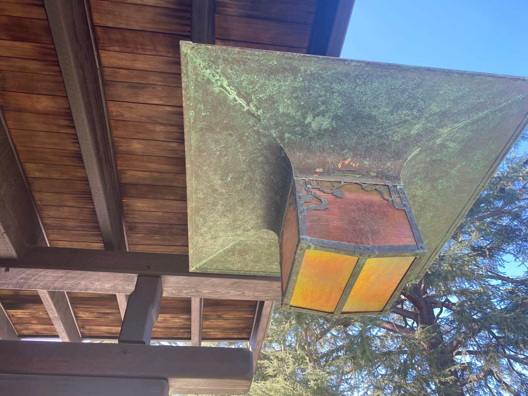 A patinated-copper lantern with amber glass mounted beneath the deep timber eaves of the Blacker House, seen from below against a blue sky.