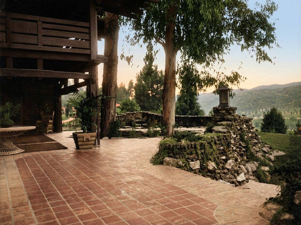 Photograph of a Greene and Greene back terrace at twilight showing brick paving extending from the house, low arroyo stone retaining walls integrated with eucalyptus trees that grow through the structure, a craftsman lantern atop a stone pier, and views over the surrounding California landscape