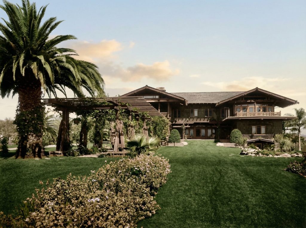 Colorized historic photograph of the gardens of the Robert R. Blacker House showing the wisteria-covered pergola walk extending from the house, mature palms framing the composition, broad lawn with curved garden beds, and the stone-faced house with its layered eaves and sleeping porches in the background