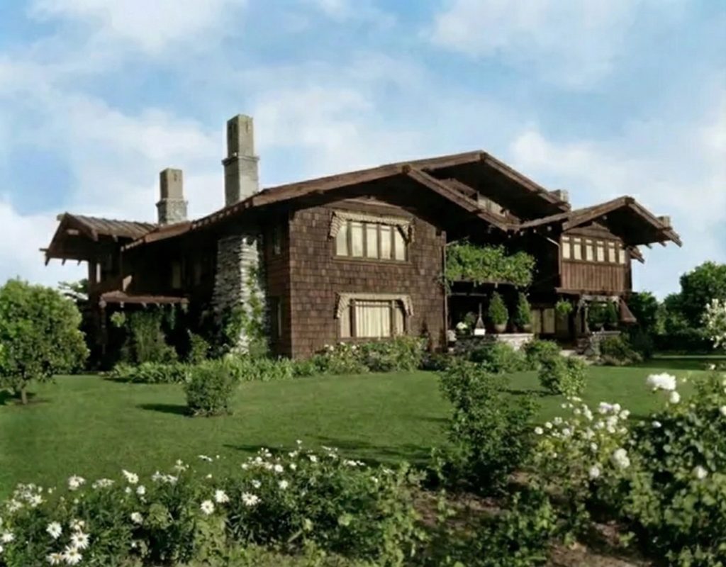 Photograph of the Mary E. Cole House by Greene and Greene showing twin stucco-capped chimneys rising above the broad gabled roof, with cedar shingle walls, deep horizontal eaves, and a foreground of white roses and lawn