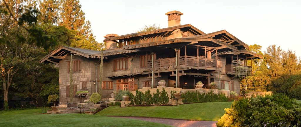 Photograph of the northeast elevation of the Gamble House by Greene and Greene at golden hour, showing the broad shingled volume, stacked sleeping porches with timber railings and posts, and the brick-and-concrete chimney caps emerging discreetly above the ridge against trees