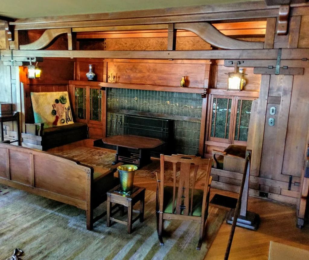 Interior view of a Greene and Greene living room showing the pronounced cloud-lift profile of the structural ceiling beams sweeping above a fireplace inglenook, with built-in cabinetry, art glass sconces, and original craftsman furniture