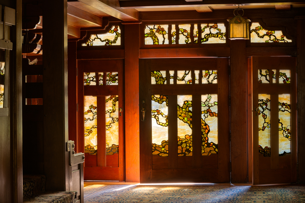 Interior view of the David B. Gamble House front doors and sidelights in full daylight, showing the leaded art glass by Emil Lange with the Japanese black pine motif rendered in amber, gold, green, and white opalescent glass panels, framed in mahogany and teak joinery