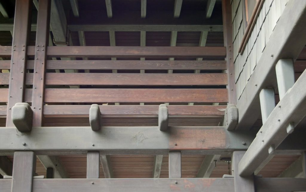 Upward view of the underside of a Greene and Greene sleeping porch showing the complex layered timber structure with expressed beam ends, stepped purlins, pegged connections, and the cedar shingle siding of the main house wall