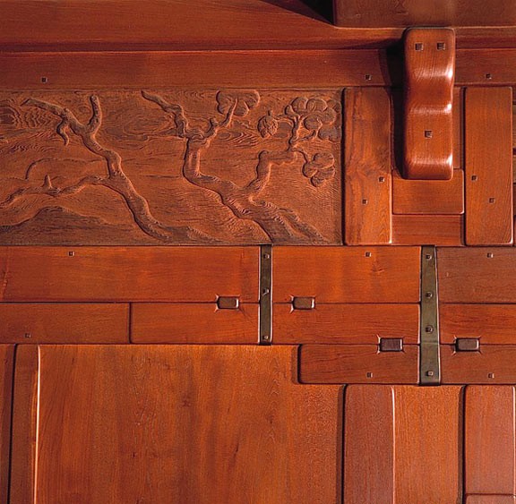 Close-up detail of Greene and Greene interior mahogany paneling showing a relief-carved landscape frieze with branching tree motif, expressed ebony pegs marking structural connections, bronze strap hardware, and the layered grammar of overlapping joinery