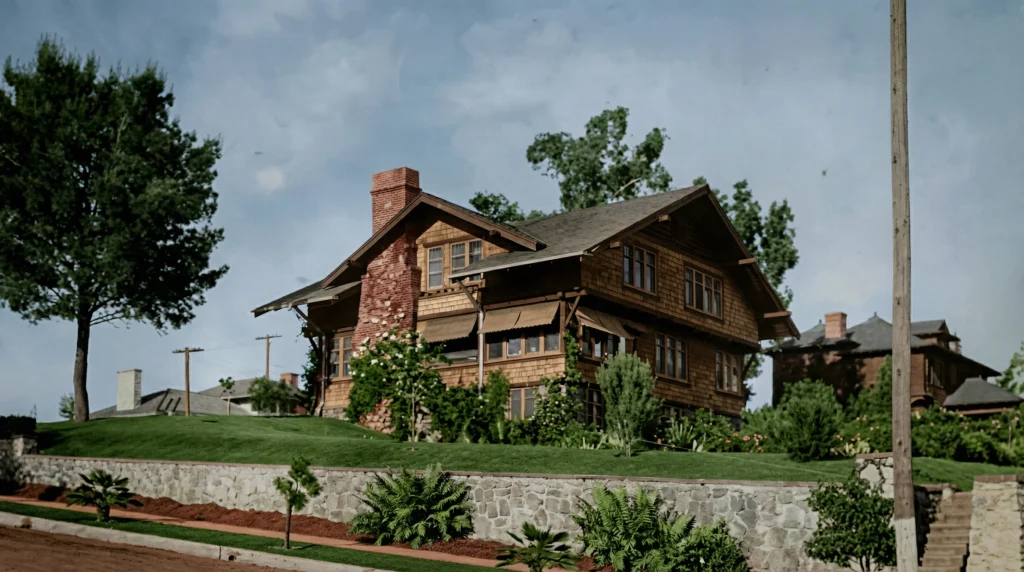 Photograph of the Lucretia Garfield House by Greene and Greene perched above a stone retaining wall, with two-story shingled walls and a tall red-brick chimney rising from the side gable, framed by mature trees against a cloudy sky
