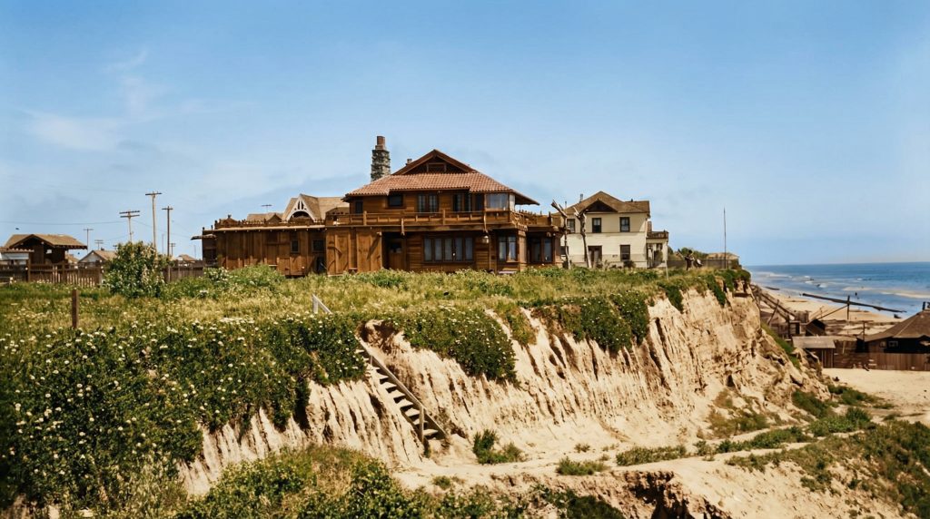 Colorized historic photograph of the Adelaide M. Tichenor House on the bluff above the Pacific Ocean in Long Beach circa 1905, showing the cedar-shingle two-story residence with deep eaves, projecting porches, a brick chimney rising at left, and an undeveloped sandy bluff in the foreground with the ocean visible at right