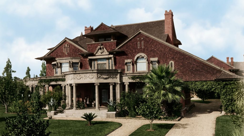 Colorized historic photograph of the James Swan House in Pasadena, designed by Charles and Henry Greene in 1898, showing the Colonial Revival composition with classical Ionic porch columns, carved swag frieze, shingled gables, and formal symmetry — an example of the brothers' early practice before their mature language emerged