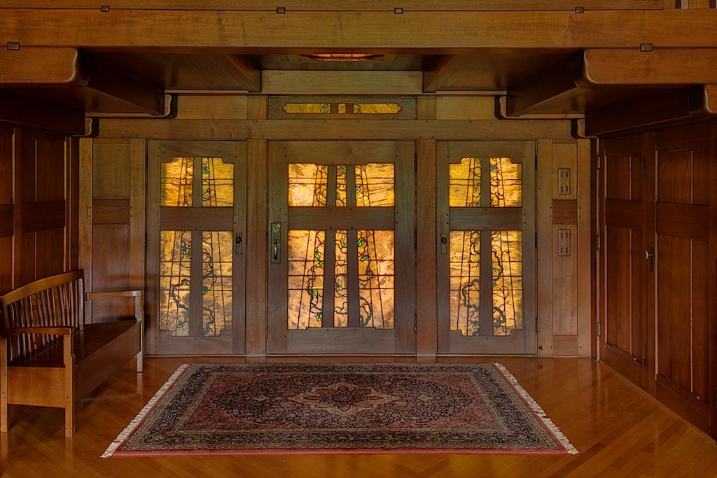 Interior view of the Robert R. Blacker House entrance hall in Pasadena, California, showing the art glass front doors and sidelights by Emil Lange rendered in warm amber and gold opalescent glass with a vine-and-branch motif, framed by mahogany and teak paneling with expressed ebony pegs, and anchored by an original Persian rug
